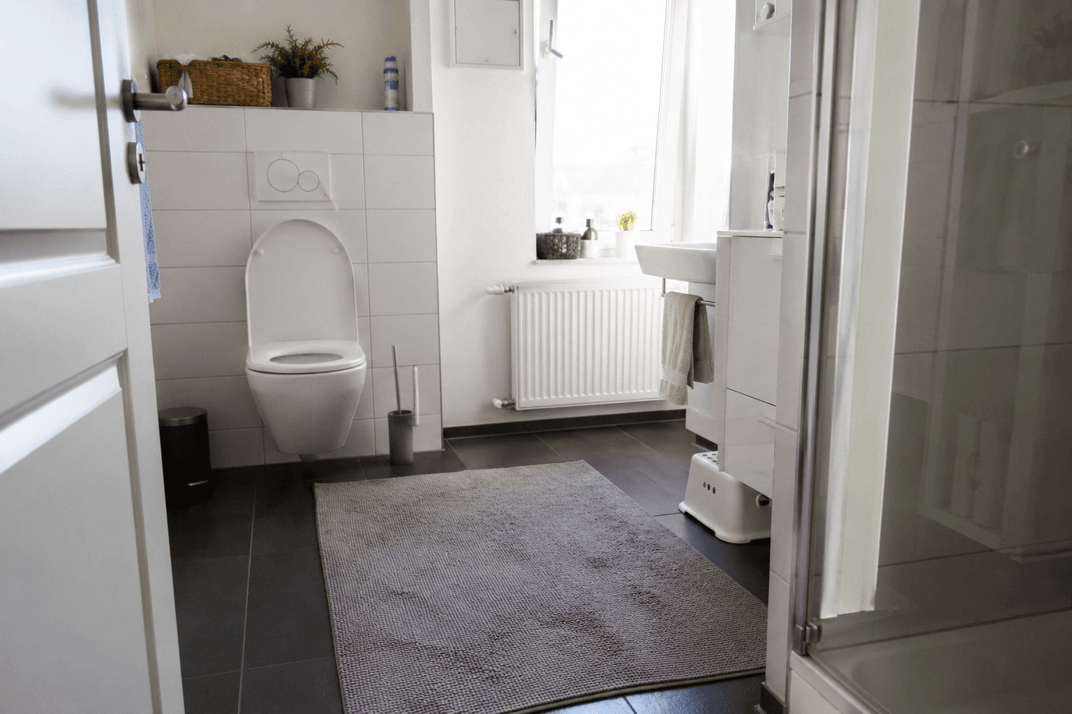 Modernes Badezimmer mit weißer Toilette, grauem Teppich, Waschbecken und Fenster über einem weißen Heizkörper.