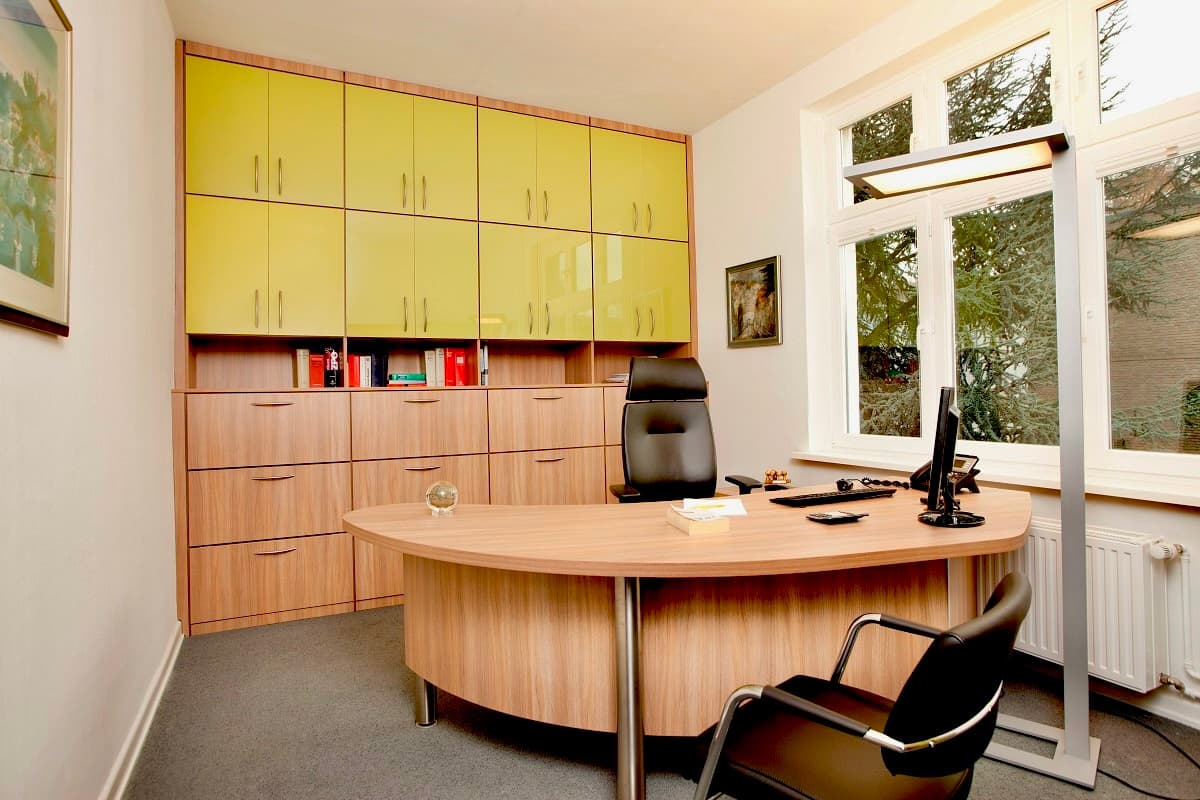Modernes Büro mit geschwungenem Holzschreibtisch, schwarzem Bürostuhl und einer großen Schrankwand mit gelben Fronten.