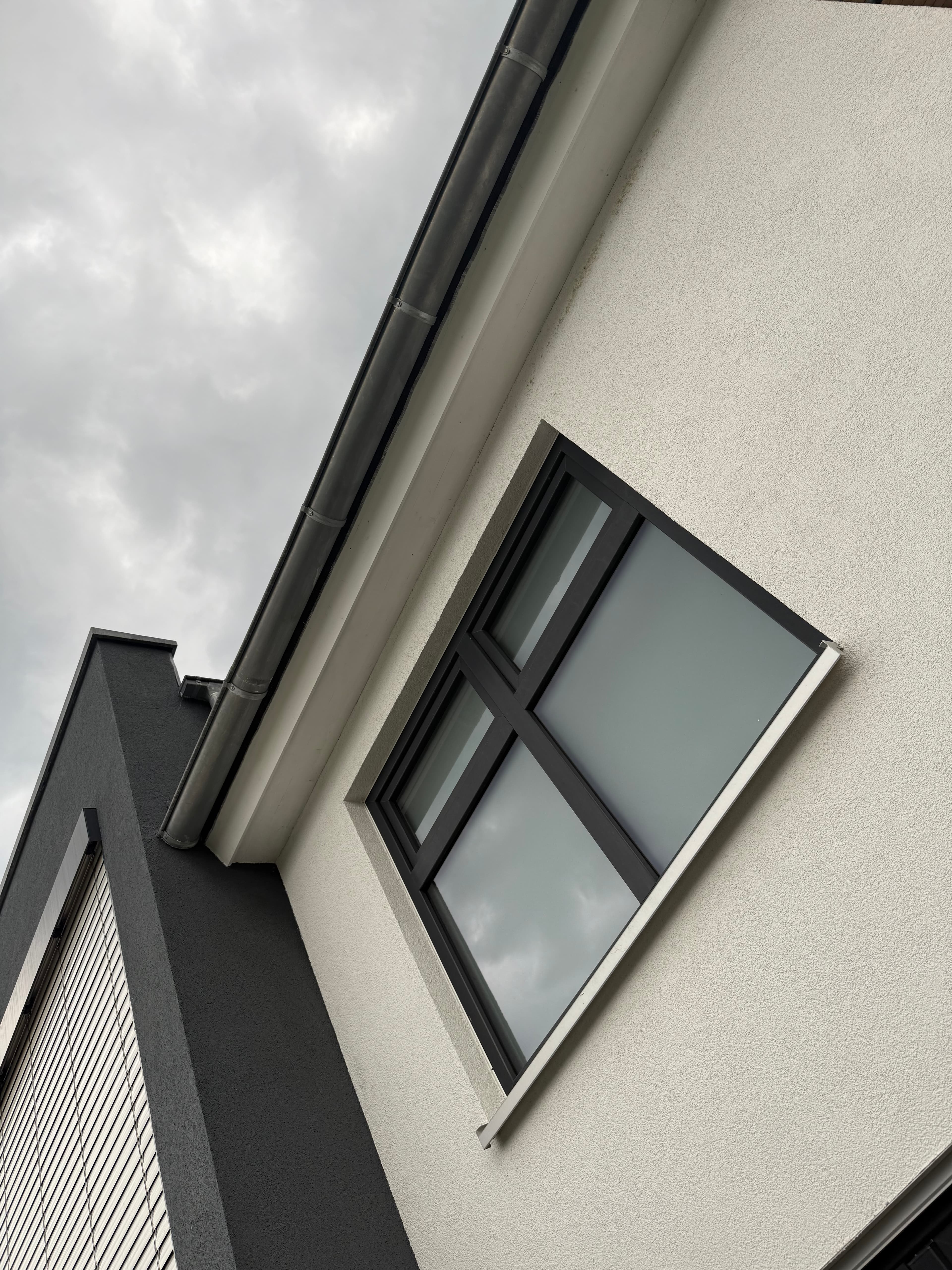 Schräge Untersicht einer modernen weißen Hauswand mit dunklem Fensterrahmen unter einem bewölkten, grauen Himmel.