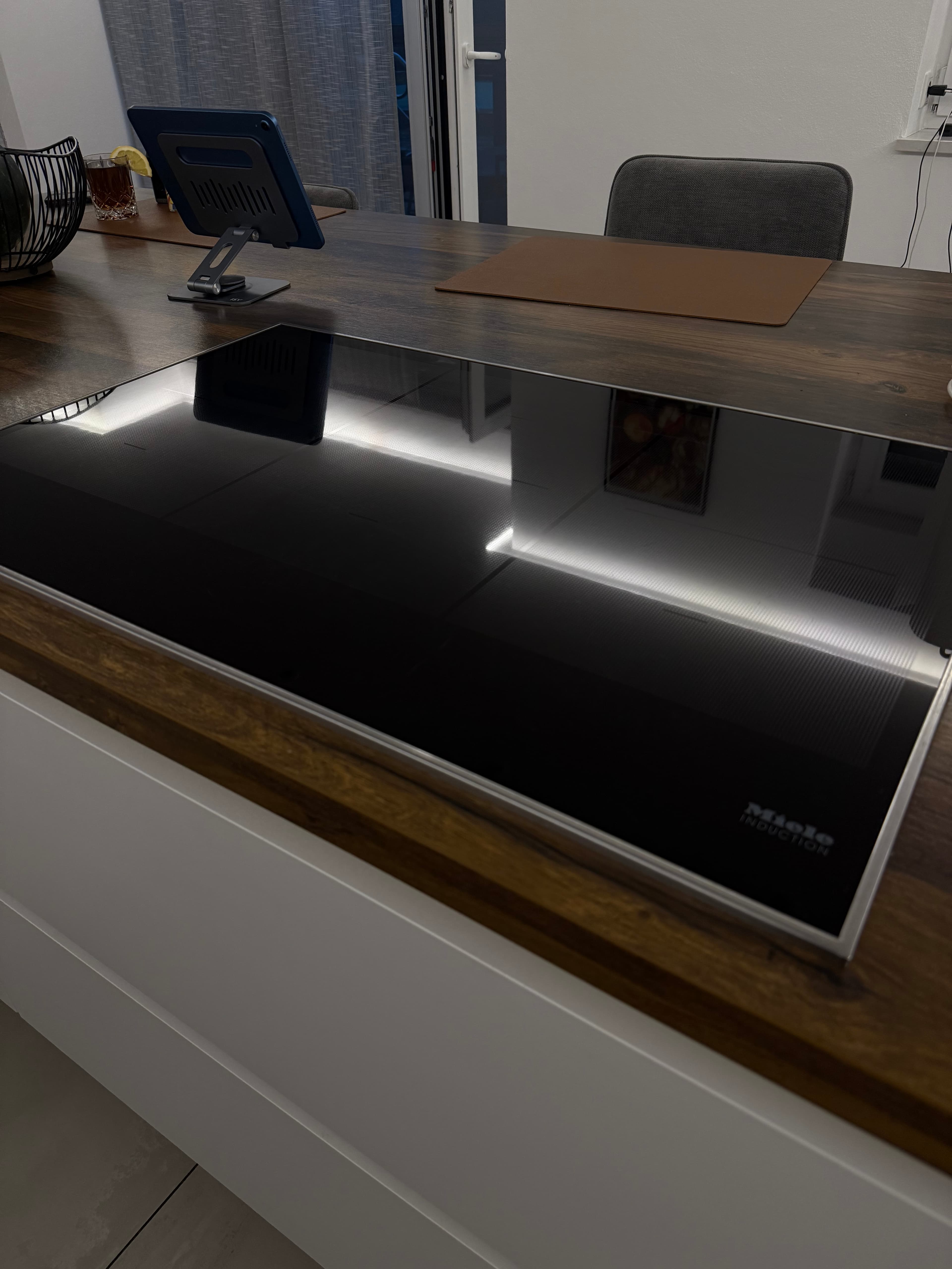 Schwarzes Miele-Induktionskochfeld auf einer dunklen Holz-Arbeitsplatte mit Spiegelungen, im Hintergrund ein Tablet und Stuhl.