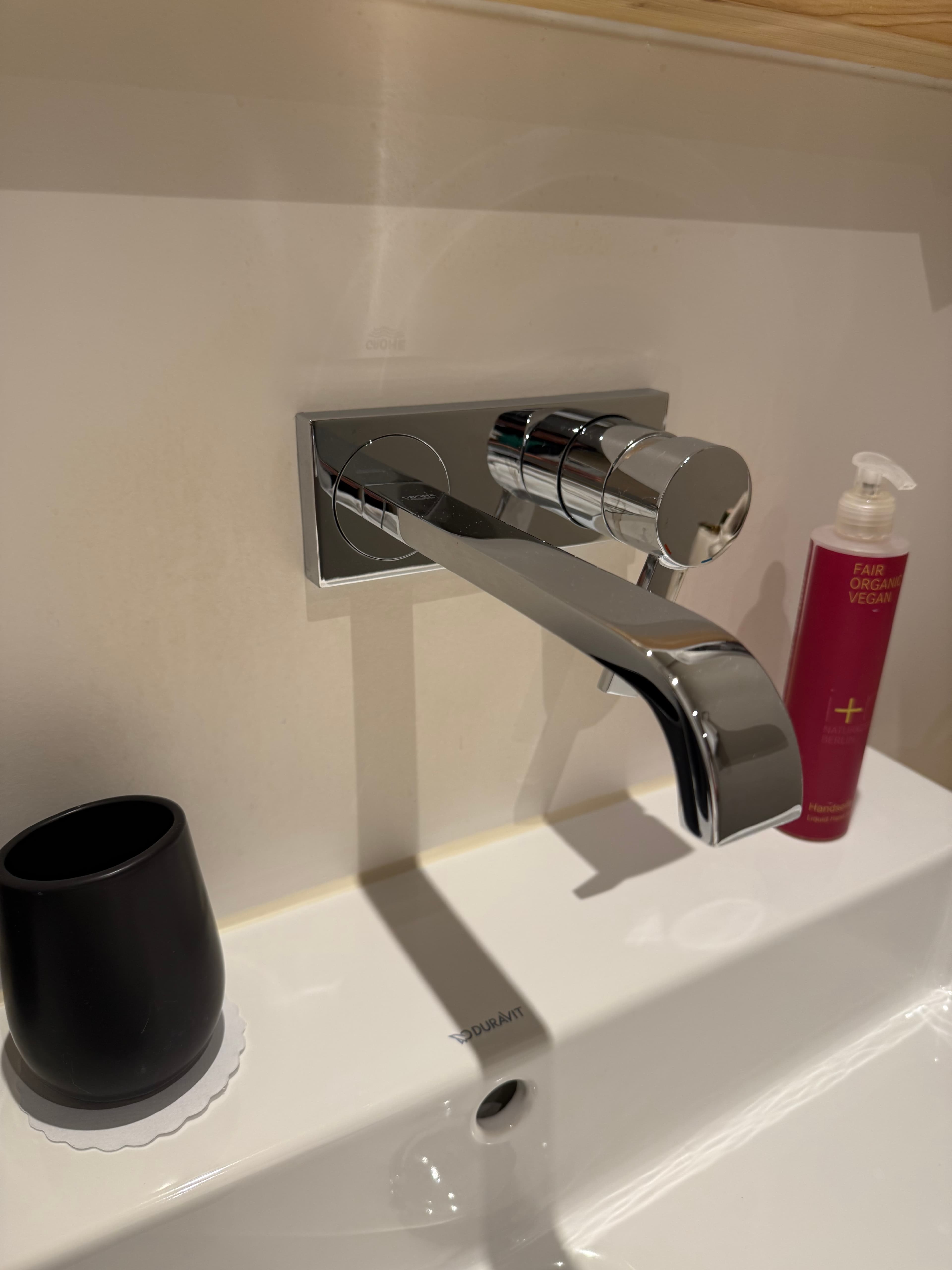 Moderne verchromte Wandarmatur über einem weißen Waschbecken mit schwarzem Becher und roter Seifenflasche.