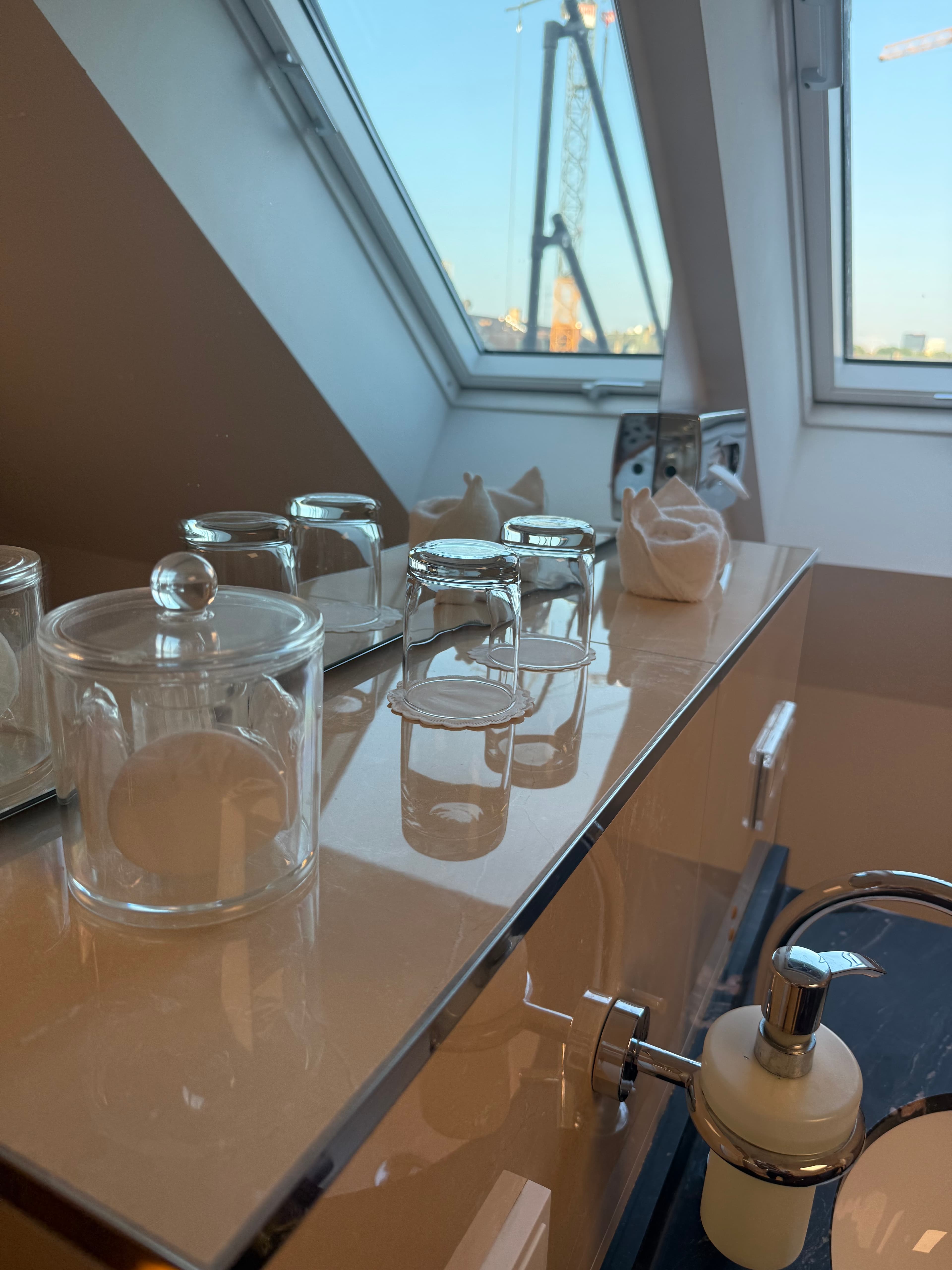 Moderner Badezimmertisch mit Glasbehältern, Trinkgläsern und einem Dachfenster, das einen Baukran im Freien zeigt.
