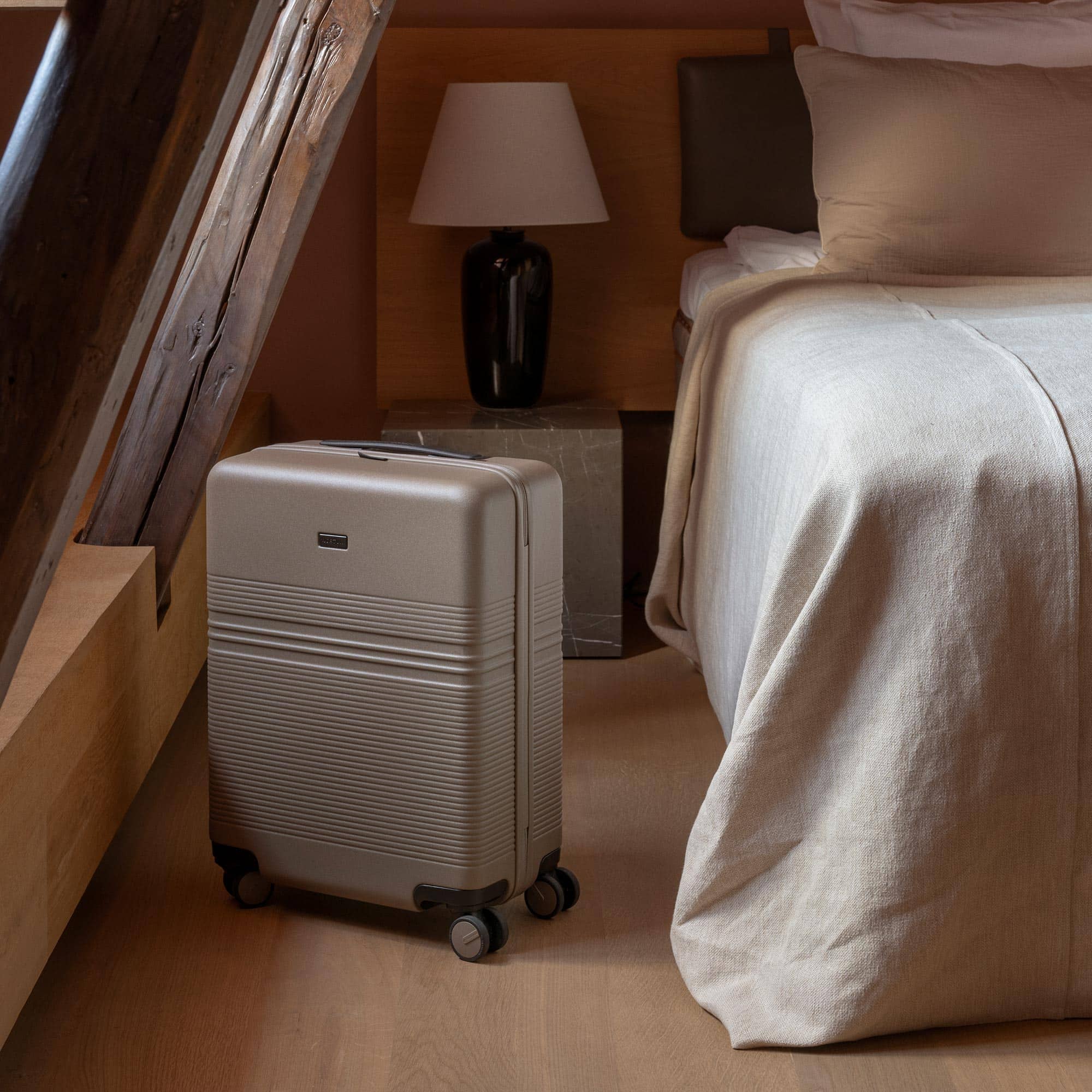 Beigefarbener Hartschalenkoffer auf Holzboden neben einem Bett in einem stilvollen Zimmer mit rustikalen Holzbalken.
