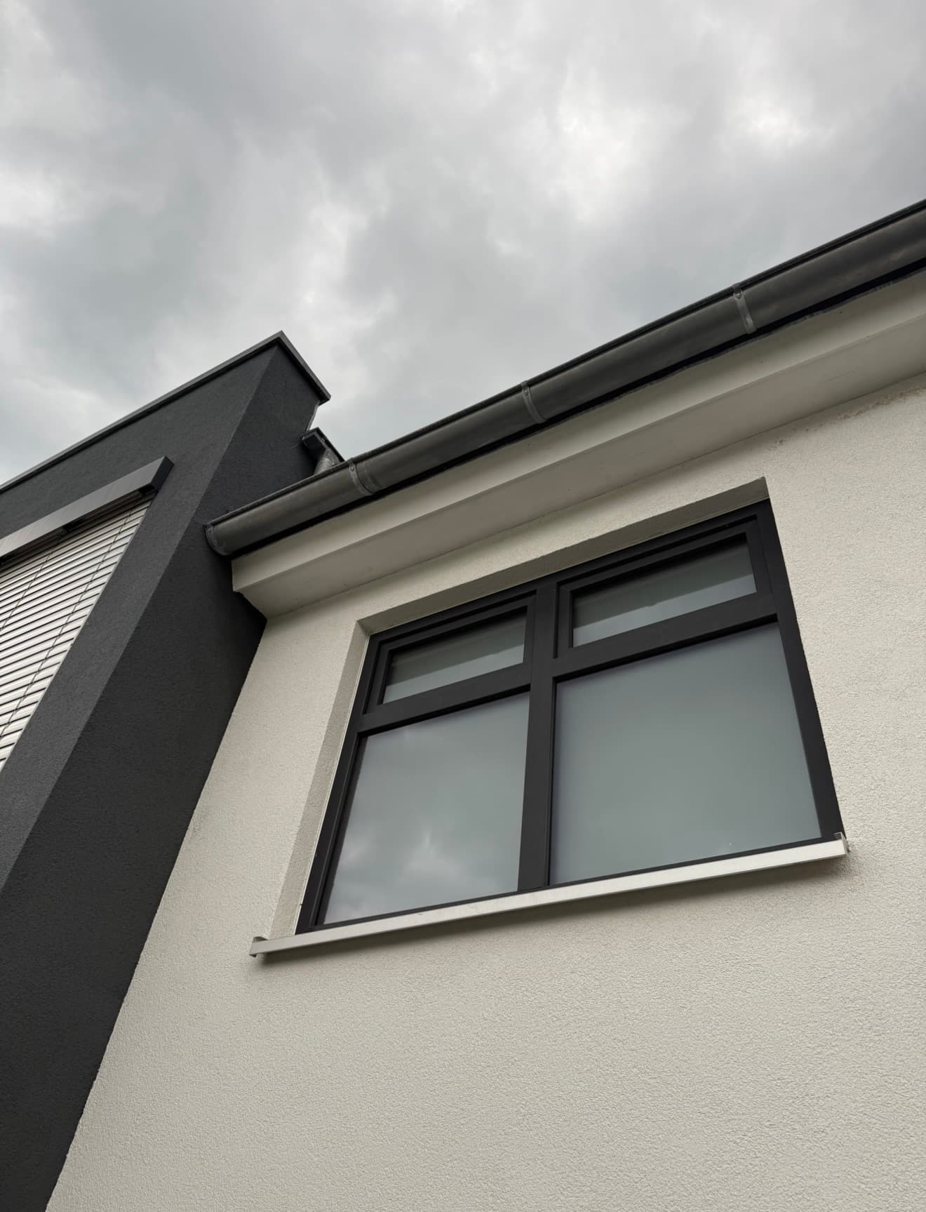 Moderne weiße Fassade mit schwarzem Fensterrahmen und dunklem architektonischem Element unter bewölktem Himmel.